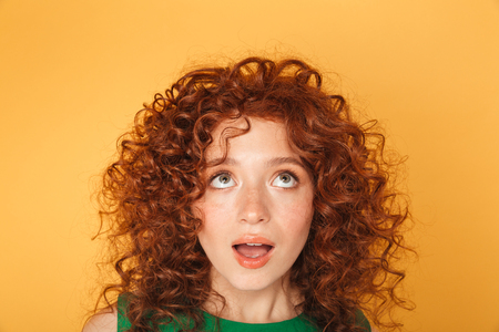 Close up of surprised young curly redhead woman looking up at copy space isolated over yellow backgroundの写真素材
