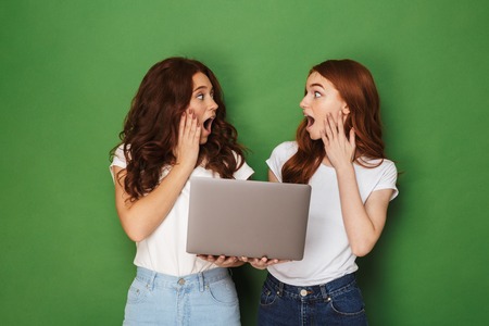 Portrait of two shocked young redhead girls holding laptop computer and looking at each other isolated over green backgroundの写真素材