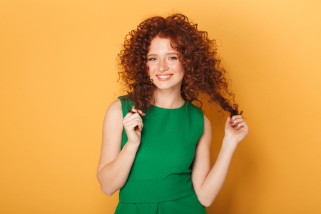 Portrait of a smiling curly redhead woman flirting and looking at camera isolated over yellow backgroundの写真素材