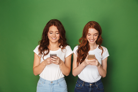 Portrait of two pretty women with ginger hair posing at camera and holding mobile phones isolated over green backgroundの写真素材