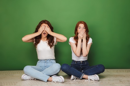 Portrait of two shocked young redhead girls sitting on a floor and cover mouth and eyes with hands isolated over green backgroundの写真素材