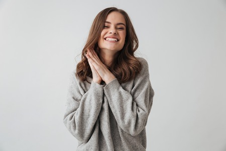 Pleased brunette woman in sweater posing with arms near face while looking at the camera over grey backgroundの写真素材