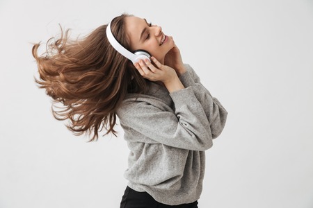 Cheerful brunette woman in sweater and headphones listening music with closed eyes and enjoys over grey backgroundの写真素材