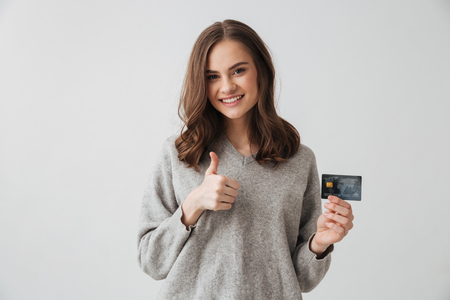 Smiling brunette woman in sweater holding credit card and showing thumb up while looking at the camera over grey backgroundの写真素材