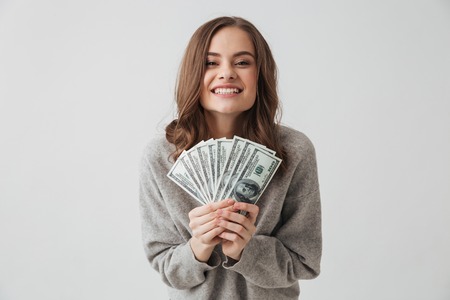 Cheerful brunette woman in sweater holding money and rejoices while looking at the camera over grey backgroundの写真素材