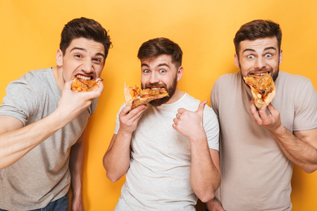 Three young satisfied men eating pizza isolated over yellow backgroundの写真素材