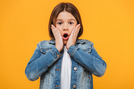Portrait of a surprised little schoolgirl looking at camera over yellow backgroundの写真素材