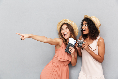 Portrait of two happy young women dressed in summer clothes holding passport with tickets and pointing at copy space over gray backgroundの写真素材