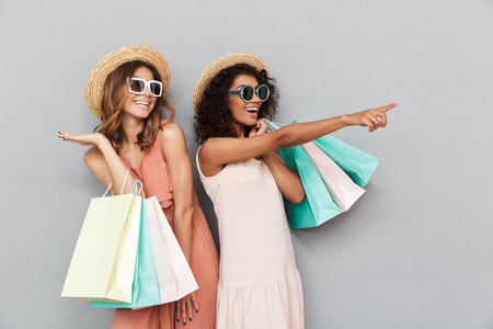 Portrait of two smiling young women dressed in summer clothes holding shopping bags and pointing at copy space isolated over gray backgroundの写真素材