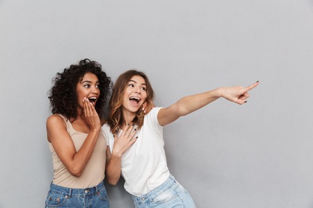 Portrait of two cheerful young women standing together and pointing away at copy space isolated over gray backgroundの写真素材