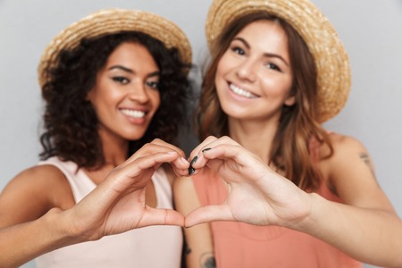 Close up portrait of two happy young women dressed in summer clothes showing heart gesture with arms isolated over gray backgroundの写真素材