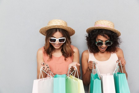 Portrait of two happy young women dressed in summer clothes looking inside shopping bags isolated over gray backgroundの写真素材