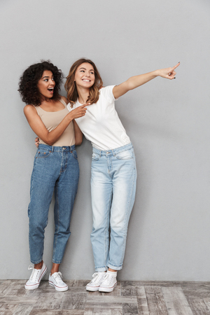 Full length portrait of two cheerful young women standing together and pointing away at copy space over gray backgroundの写真素材
