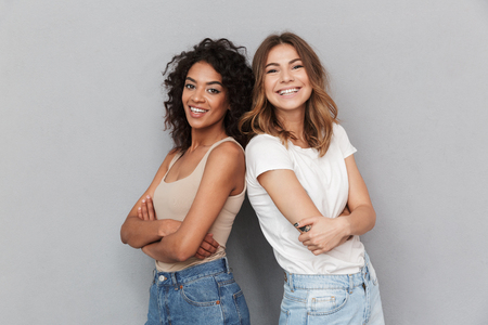 Portrait of two cheerful young women standing together and looking at camera isolated over gray backgroundの写真素材