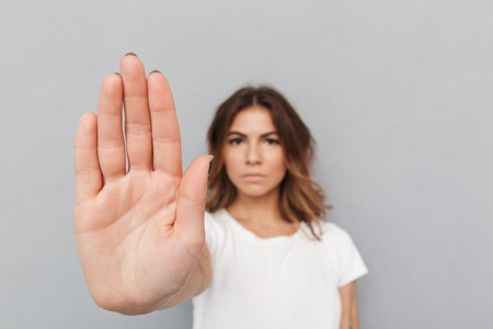 Portrait of a serious young woman showing stop gesture with her palm over gray backgroundの写真素材