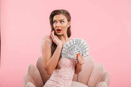 Portrait of fancy caucasian woman in dress holding fan of money while sitting in armchair isolated over pink backgroundの写真素材