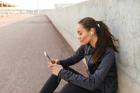 Image of serious young asian sports woman sitting outdoors listening music using mobile phone.の写真素材
