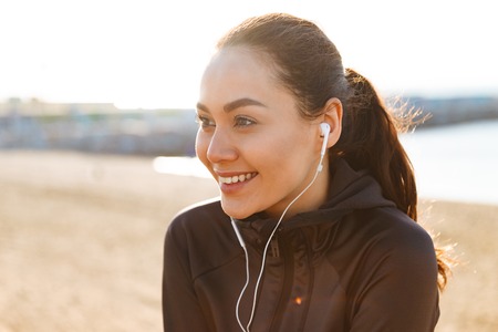 Photo of cheerful young asian sports woman sitting outdoors listening music looking aside.の写真素材