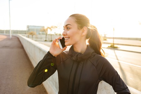 Image of happy young asian sports woman walking outdoors talking by mobile phone.の写真素材