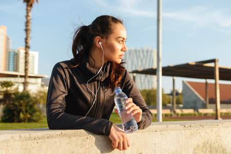 Image of beautiful young asian sports woman listening music with earphones drinking water outdoors.の写真素材
