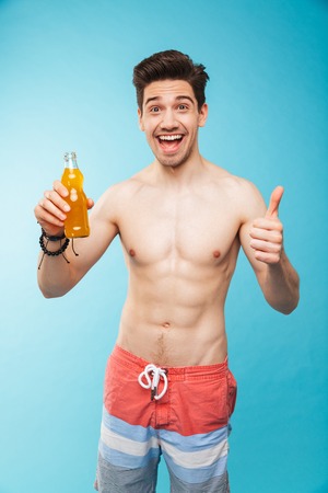 Portrait if a cheerful shirtless man showing beer bottle and giving thumbs up over blue backgroundの写真素材