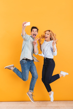 Full length photo of happy man and woman taking selfie photo on mobile phone while jumping and showing victory sign isolated over yellow backgroundの写真素材