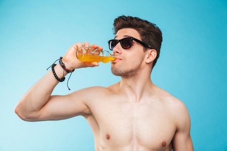 Close up portrait of a shirtless young man in sunglasses drinking beer from a bottle over blue backgroundの写真素材