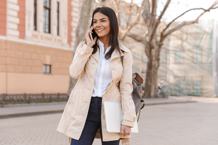 Smiling young woman dressed in autumn coat talking on mobile phone while carrying laptop and walking on a city streetの写真素材