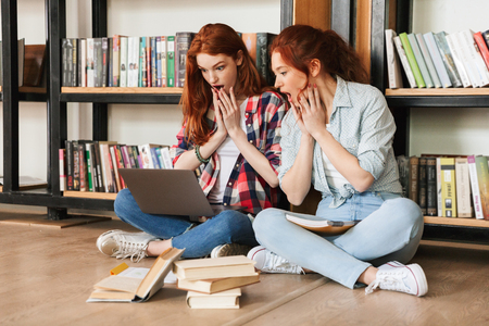 Two shocked teenage girls sitting on a floor at the bookshelf in a library and doing homework with laptop computerの写真素材