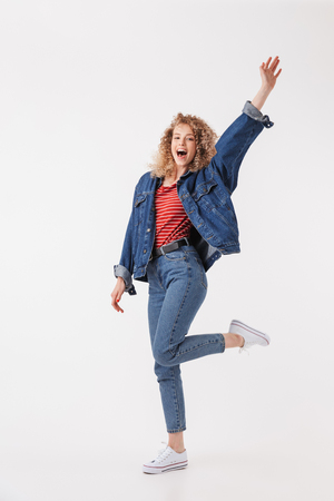 Full length image of Cheerful blonde curly woman in denim clothes rejoices and looking at the camera over grey backgroundの写真素材