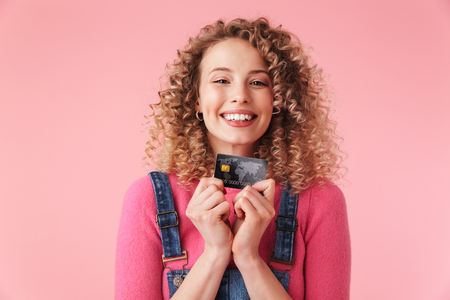 Portrait of delighted young girl with curly hair holding credit card isolated over pink backgroundの写真素材