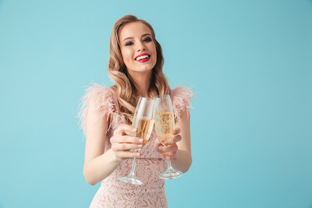 Photo of beautiful woman 20s wearing pink dress with makeup smiling and holding two glasses of champagne isolated over blue backgroundの写真素材
