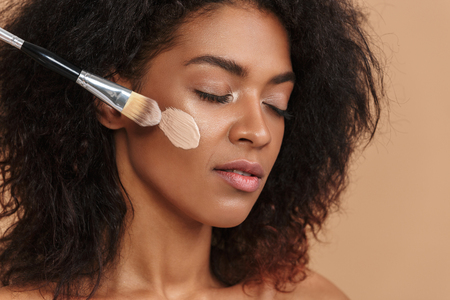 Close up image of Young pretty african woman using cosemetic brush with closed eyes over beige backgroundの写真素材