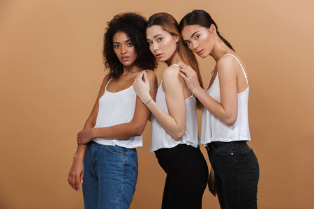 Image of three beautiful women of different nation: caucasian, african american and asian girls in basic clothing standing together isolated over beige backgroundの写真素材