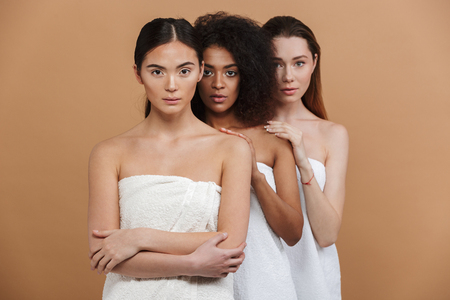 Three young multiethnic women wearing in towels posing together and looking at the camera over beige backgroundの写真素材
