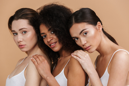 Image closeup of three gorgeous women of different nation: caucasian, african american and asian girls in basic clothing standing together isolated over beige backgroundの写真素材