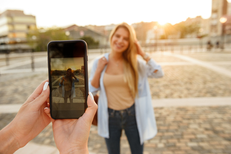 Photo of woman holding mobile phone and photographing her friend blonde lady outdoors.の写真素材