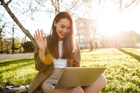 Image of amazing beautiful woman sitting on grass outdoors using laptop computer waving to friends.の写真素材