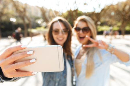 Image of two amazing smiling women friends walking outdoors make selfie by mobile phone.の写真素材