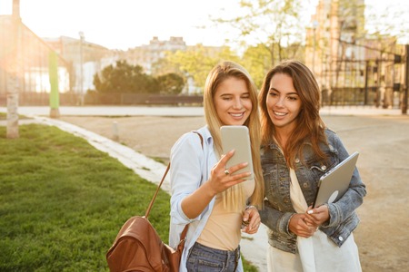 Image of happy two friends women in park outdoors holding laptop computer and using mobile phone...