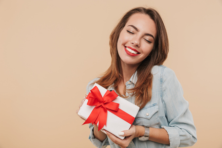 Portrait of a satisfied young girl holding present box isolated over beige backgroundの写真素材