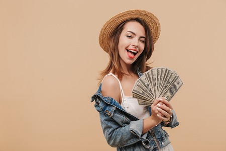 Portrait of a smiling young girl in summer clothes showing money banknotes and winking isolated over beige backgroundの写真素材