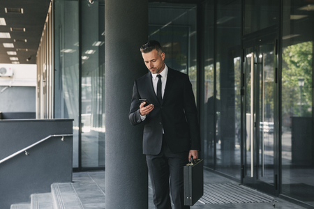 Portrait of a charming young businessman dressed in suit with briefcase walking outside a glass building and using mobile phoneの写真素材