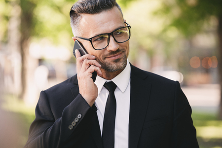 Portrait of a happy businessman dressed in suit walking outside and using mobile phoneの写真素材