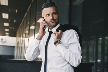 Portrait of a focused young businessman dressed in formal clothes standing outside a glass building with jacket over his shoulder and talking on mobile phoneの写真素材