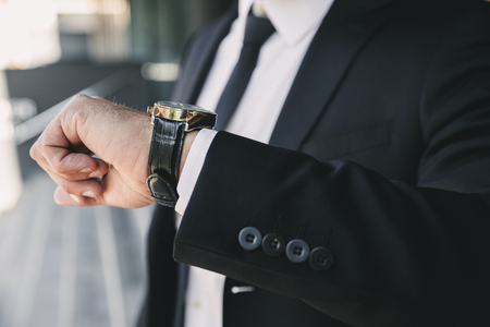 Close up of a businessman dressed in suit standing outside a glass building and looking at his wrist watchの写真素材