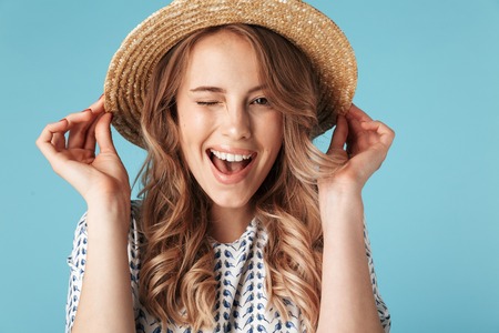 Cheerful Blonde woman in dress holding hat while winks and looking at the camera over blue backgroundの写真素材