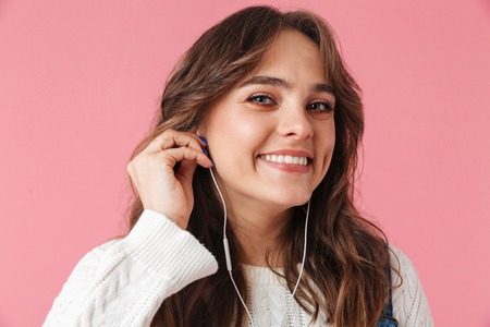 Close up of a smiling young girl listening to music with earphones isolated over pink background