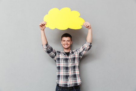 Portrait of a smiling young man in plaid shirt showing blank screen speech bubble over gray...