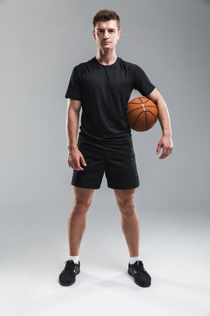 Full length portrait of a concentrated young sportsman standing with a basketball isolated over gray backgroundの写真素材